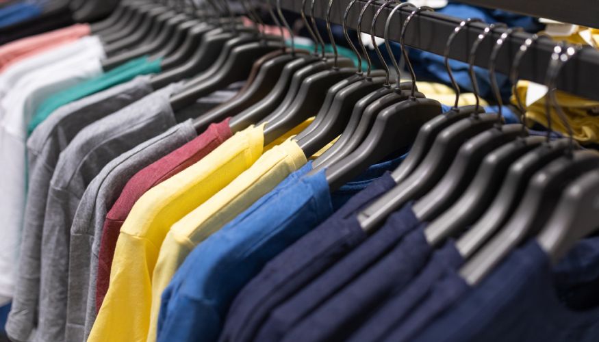 A variety of colorful t-shirts hanging on black hangers on a store rack, featuring shades of yellow, blue, gray, and red.