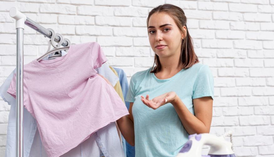 A woman prepares to wash a pink t-shirt, standing by a clothes rack against a white brick wall.